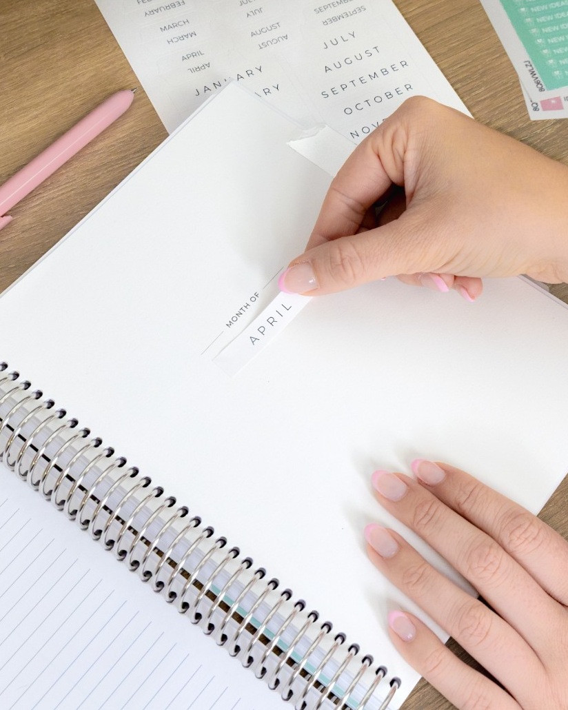Planner on a table with hands placing a label on an undated planner Planner on a table with hands placing a label on an undated planner